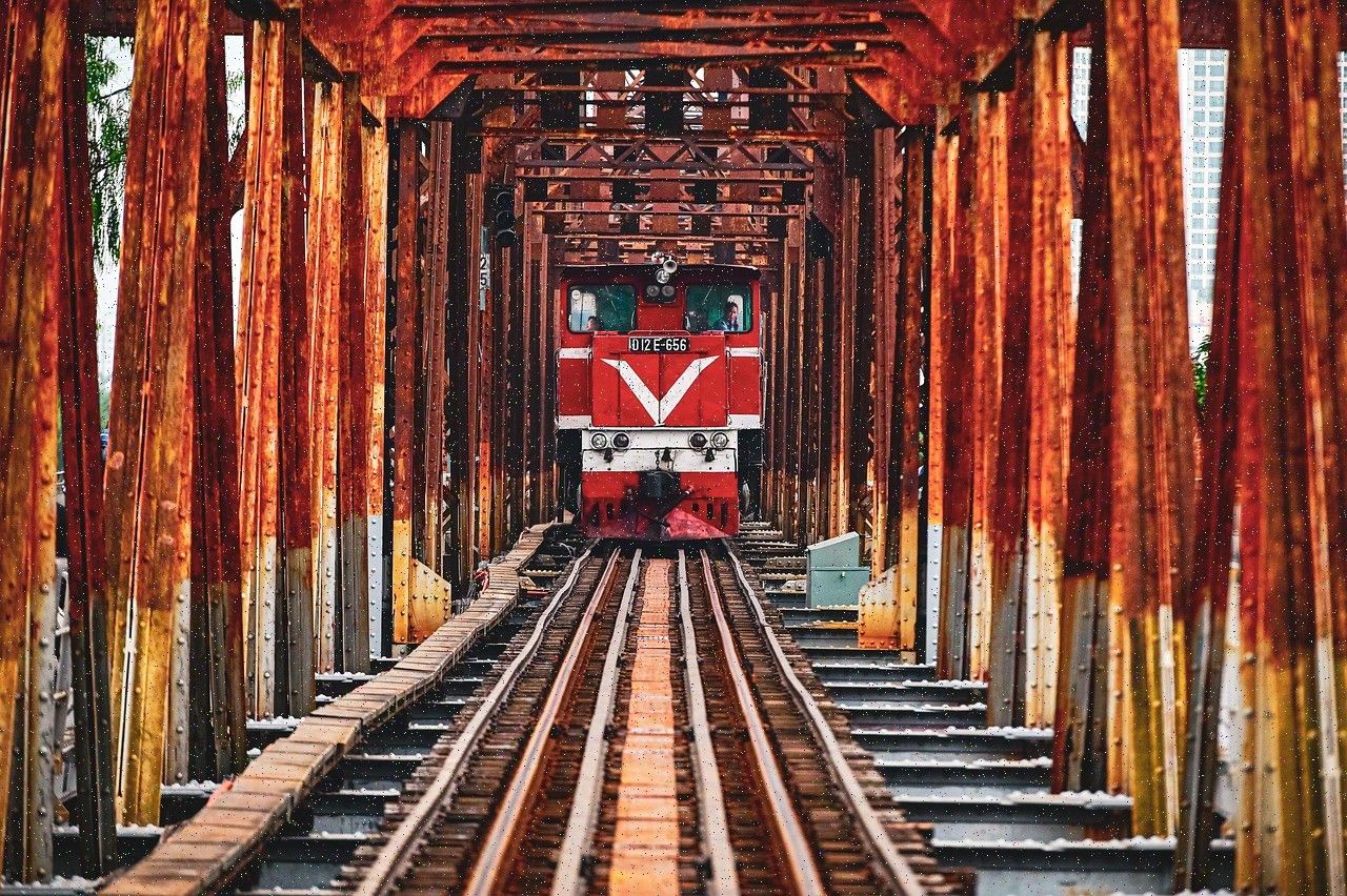 A red locomotive framed by the rusted steel trusses of a railway bridge, seen head on.