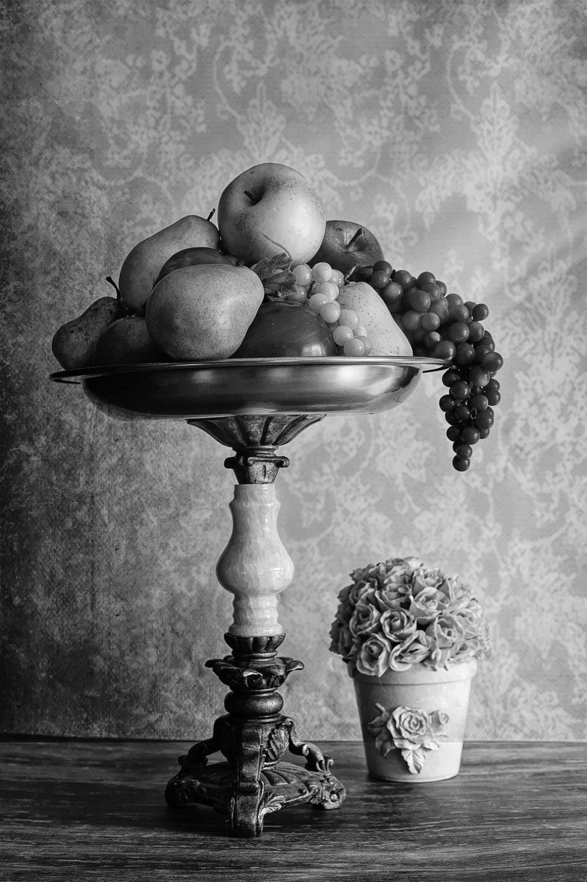 Black and white still life of fruit on a pedestal dish beside a small pot of roses.